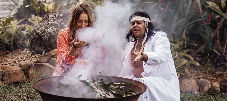 Mindy Woods sitting next to an aboriginal woman and breathing in smoke from leaves