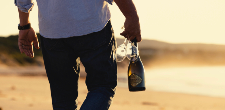 A man walking on a beach at sunset with a bottle of Arras sparkling and glasses in his hand.